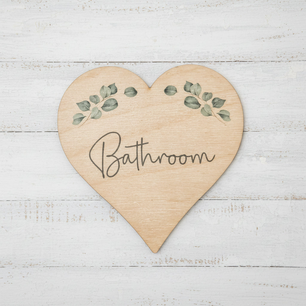 A heart-shaped wooden bathroom sign with a natural light wood grain finish, featuring green eucalyptus leaf illustrations and the word 'Bathroom' written in black cursive script, shown on a plain white background.
