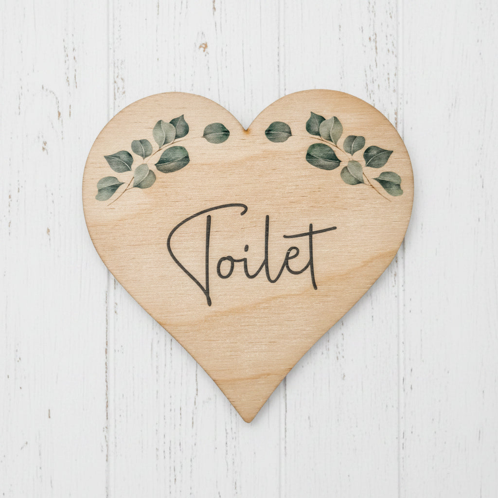 A heart-shaped wooden sign with the word 'Toilet' written in black script, decorated with green leafy branches at the top, shown on a plain white background.