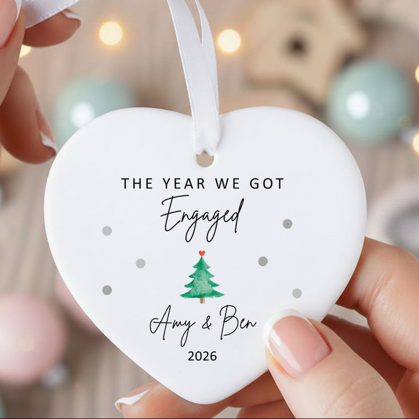 A person holds a white heart-shaped ceramic Christmas ornament with a white ribbon. The ornament is printed with the text 'THE YEAR WE GOT Engaged 2026' and a small green Christmas tree with a red heart on it.
