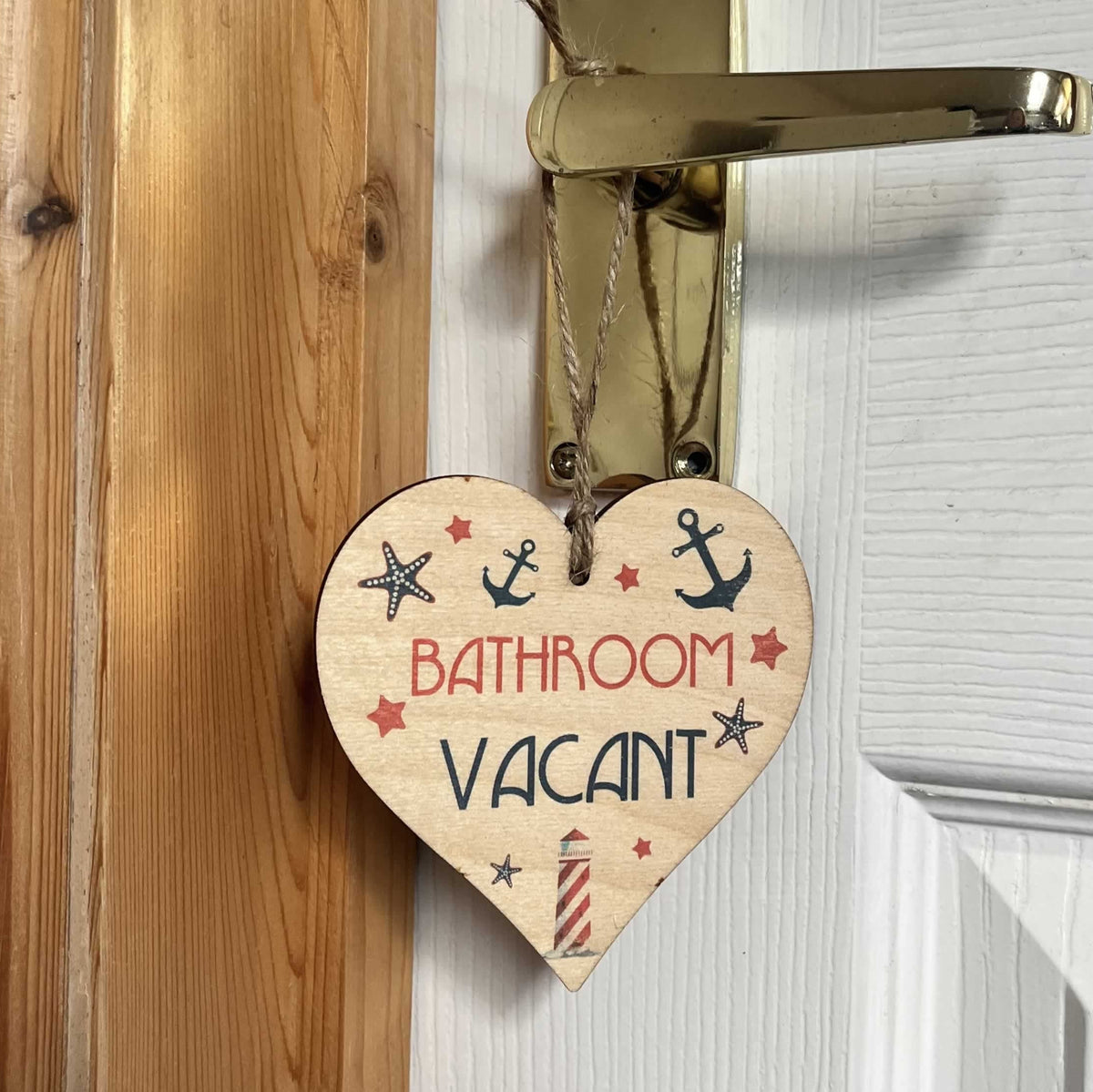 A heart-shaped wooden bathroom door hanger with the words 'BATHROOM VACANT' in red and navy lettering, decorated with small anchor, star, and lighthouse illustrations, hanging from a brass doorknob next to a white paneled door.