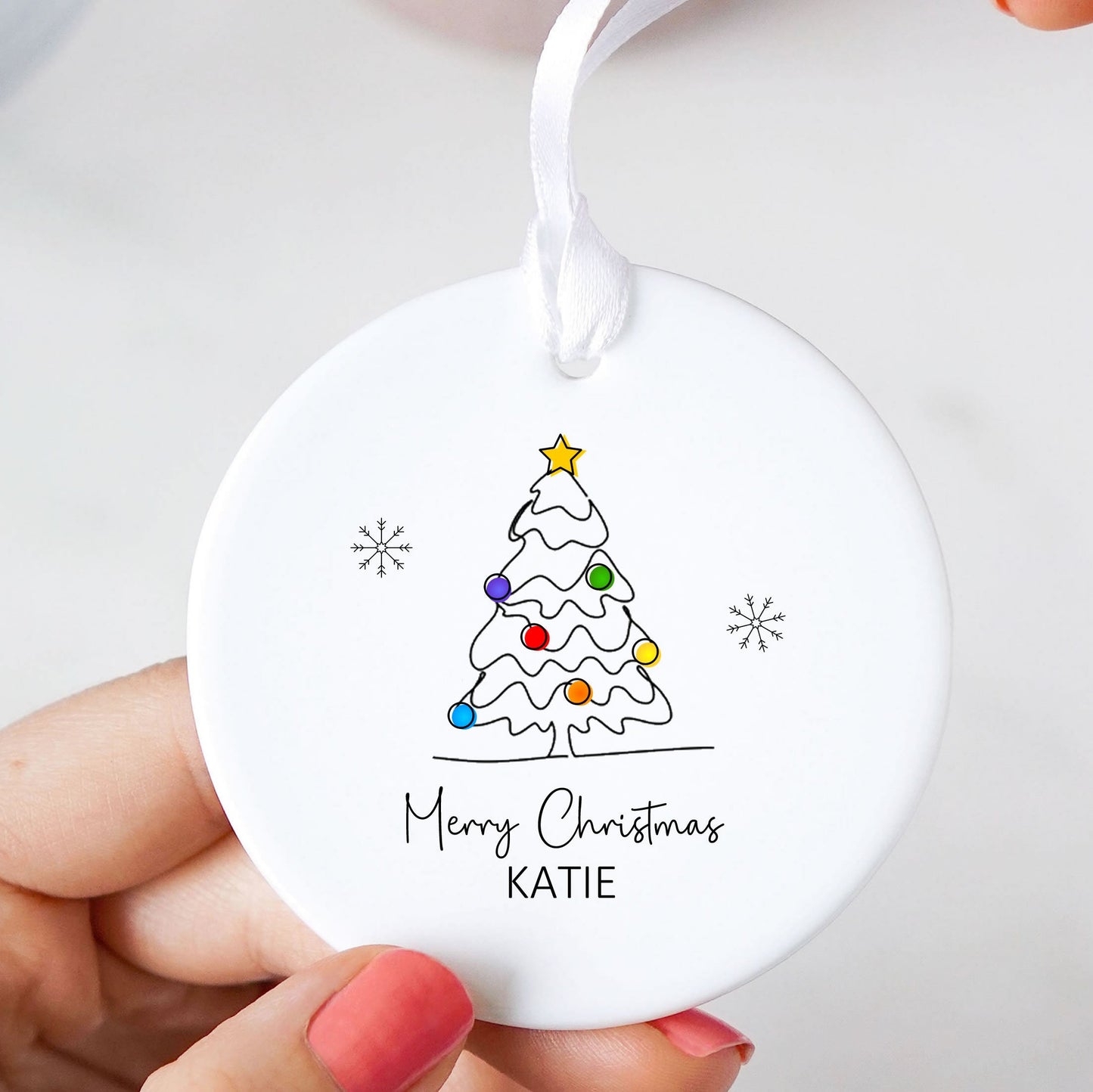 A round white ceramic Christmas ornament being held by hands with pink nail polish. The ornament features a simple black line drawing of a decorated Christmas tree with colorful ornaments, a gold star, and the text 'Merry Christmas KATIE' printed below.