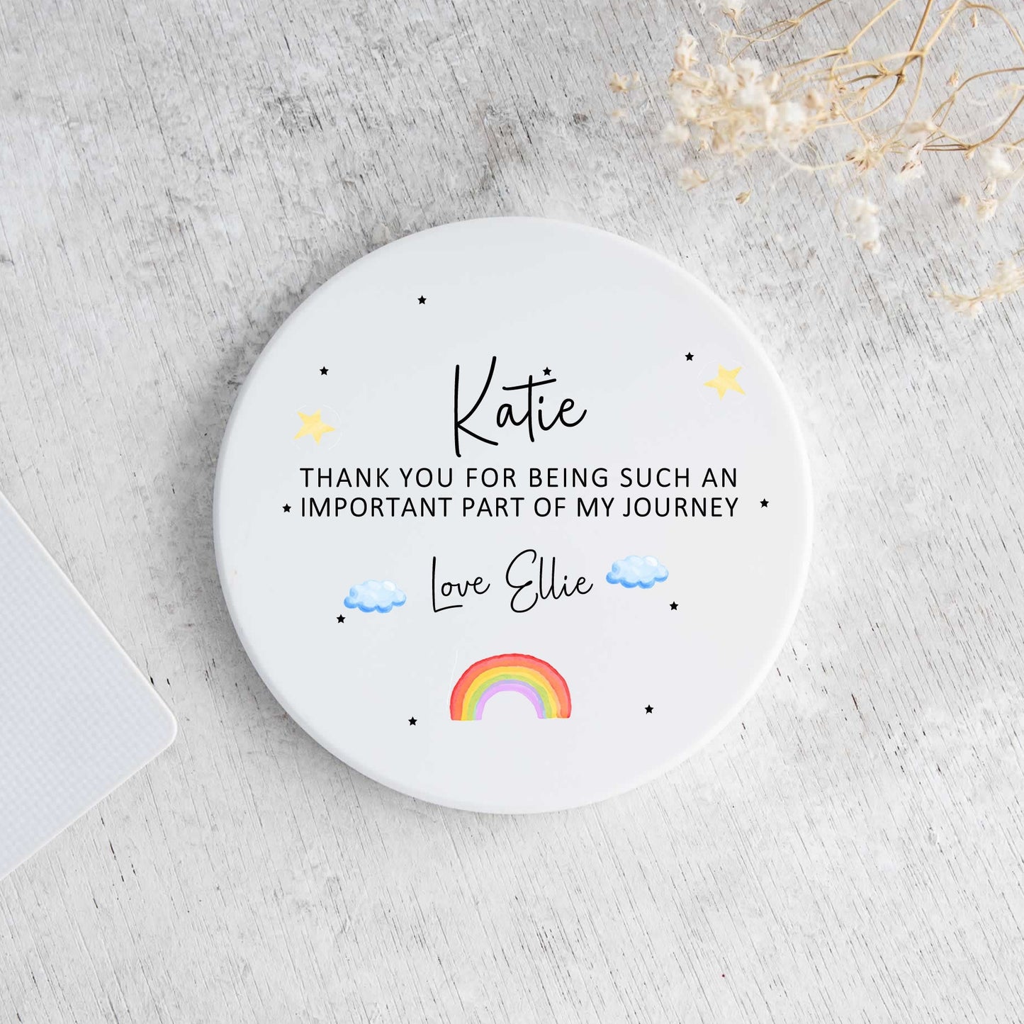 A round white coaster with personalized black text and small colorful illustrations, photographed from above on a light gray textured surface with part of a laptop and dried flowers visible.