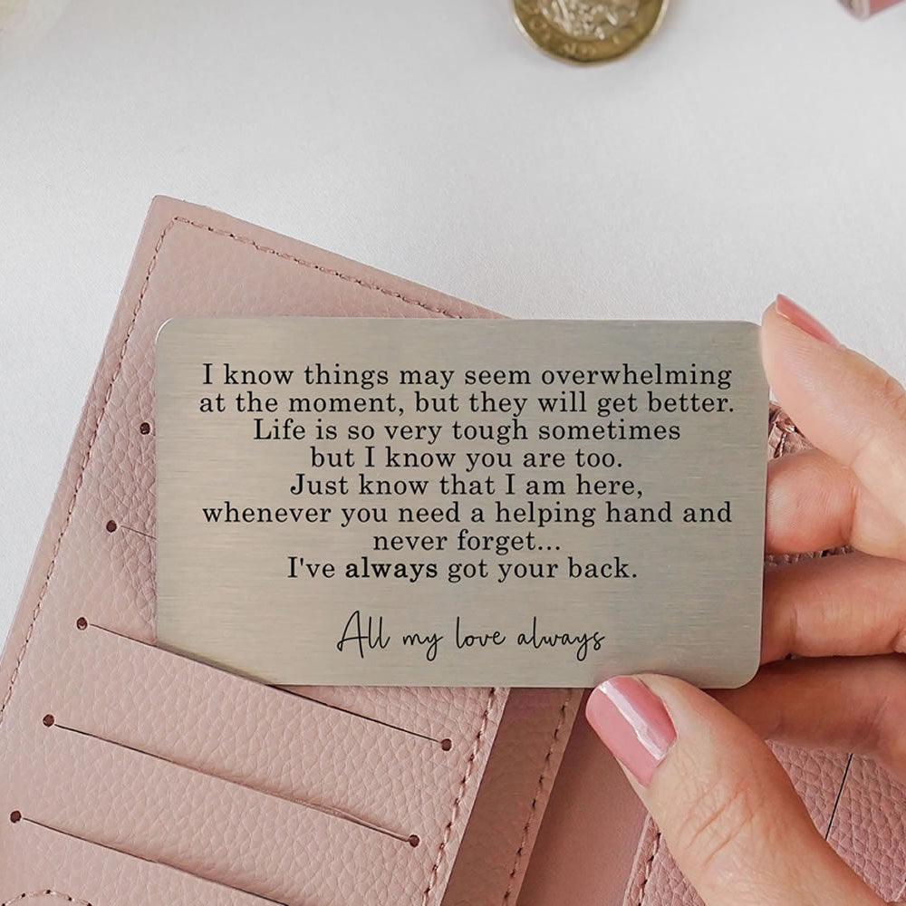 Hand holding a metal wallet card with an engraved supportive message, placed above a pink wallet.