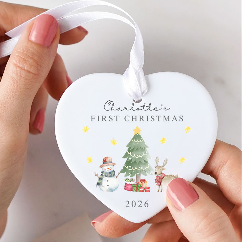 A person holds a white heart-shaped Christmas ornament with a white ribbon loop. The ornament is personalized with the text 'Charlotte's FIRST CHRISTMAS' and the year '2026', decorated with a watercolor-style illustration of a Christmas tree, snowman, gifts, and a reindeer with small yellow stars.