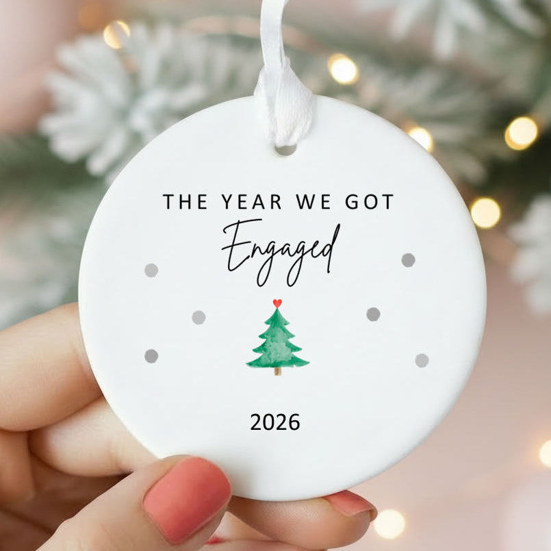 A round white ceramic Christmas ornament held by two hands, featuring the printed text 'THE YEAR WE GOT Engaged 2026' with a small green Christmas tree illustration. The background is a soft-focus holiday scene with lights and pine branches.