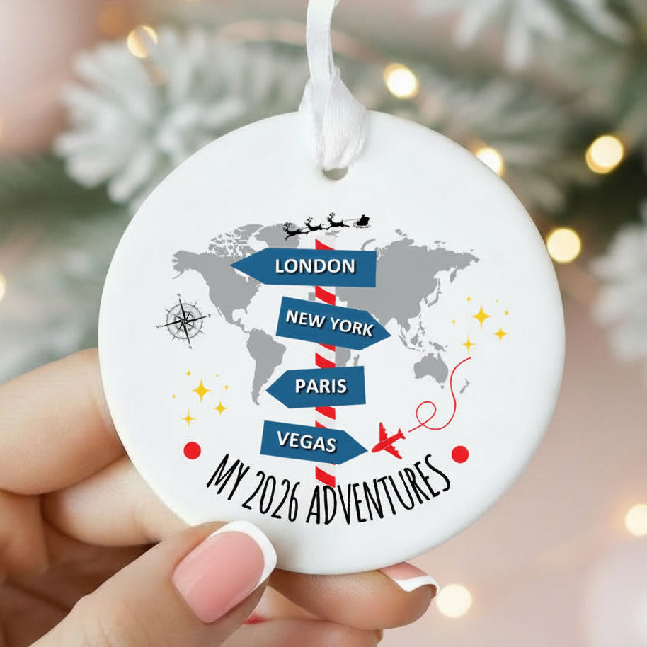 A round white ceramic Christmas ornament held by hands, featuring a gray world map with blue directional arrows pointing to London, New York, Paris, and Vegas, plus the text 'MY 2026 ADVENTURES'.