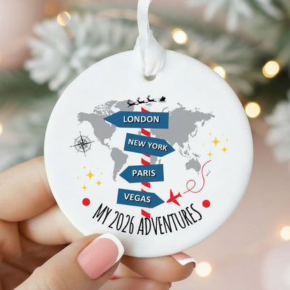 A round white ceramic Christmas ornament held by hands, featuring a gray world map with blue directional arrows pointing to London, New York, Paris, and Vegas, plus the text 'MY 2026 ADVENTURES'.