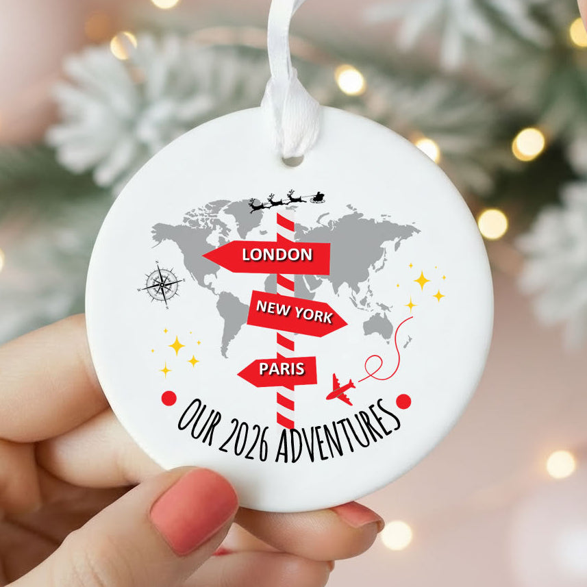 A round white Christmas ornament held by hands, featuring a gray world map, red directional arrows for London, New York, and Paris, and the text 'OUR 2026 ADVENTURES' against a softly blurred festive background.