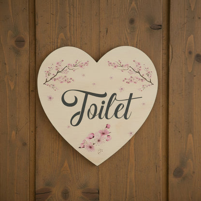 Heart-shaped wooden sign with pink cherry blossom illustrations and the word 'Toilet' in black script, shown on a plain white background.