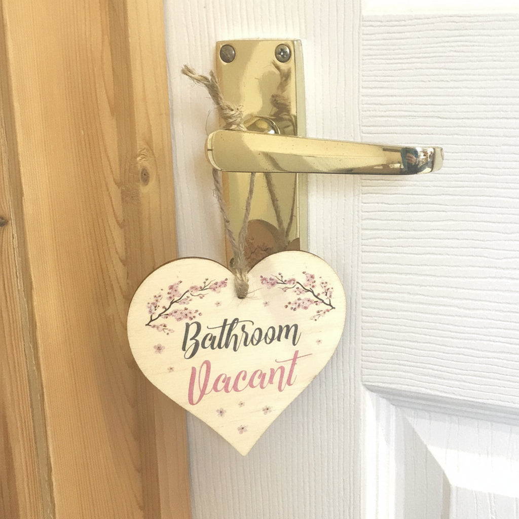 A heart-shaped wooden door hanger with the words 'Bathroom Vacant' in black and pink script, decorated with pink floral branches and small pink stars, hanging from a gold door handle on a white paneled door next to a wooden doorframe.