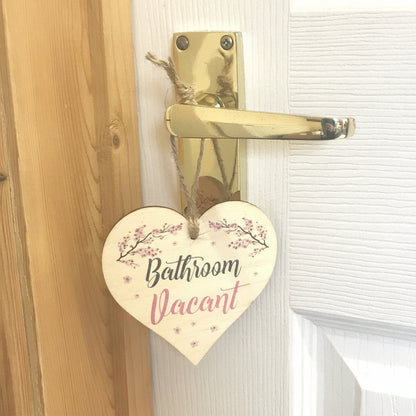 A heart-shaped wooden door hanger with the words 'Bathroom Vacant' in black and pink script, decorated with pink floral branches and small pink stars, hanging from a gold door handle on a white paneled door next to a wooden doorframe.