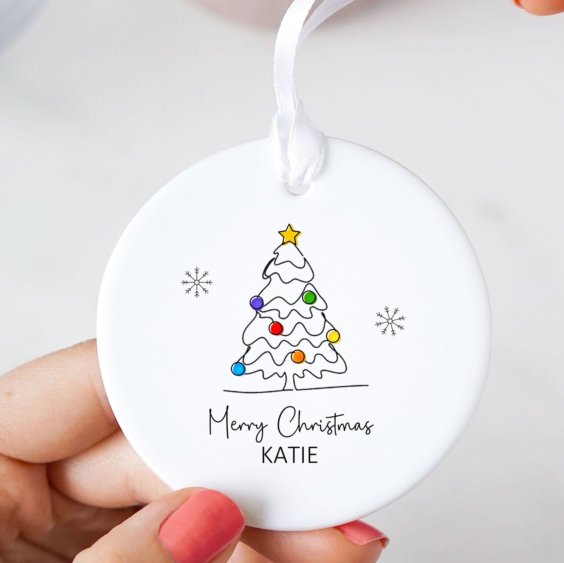 A round white ceramic Christmas ornament being held by hands with pink nail polish. The ornament features a simple black line drawing of a decorated Christmas tree with colorful ornaments, a gold star, and the text 'Merry Christmas KATIE' printed below.