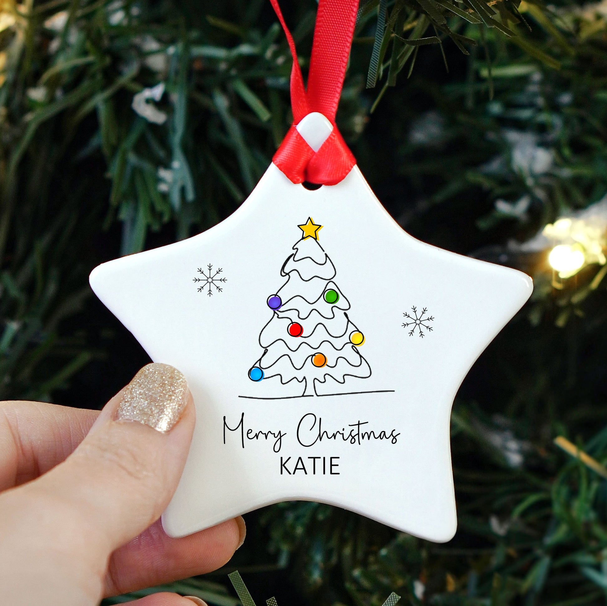 A hand with glittery nail polish holds a white star-shaped ceramic Christmas ornament featuring a hand-painted tree and the text 'Merry Christmas KATIE' in front of a blurred Christmas tree with lights.