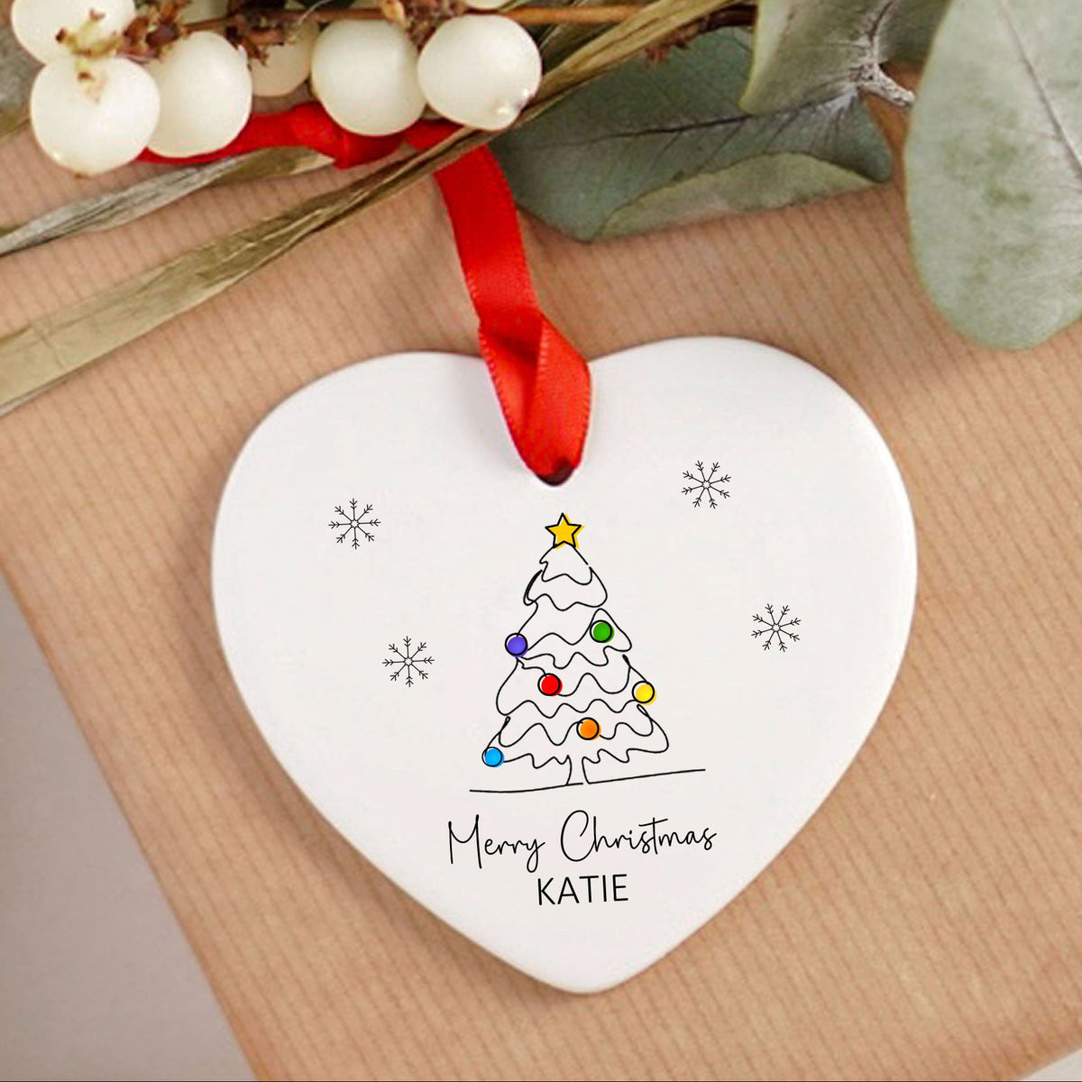 A white heart-shaped ceramic Christmas ornament with a red ribbon, featuring a hand-painted Christmas tree and the personalized text 'Merry Christmas KATIE', displayed on a brown kraft paper gift box with eucalyptus leaves and white berries.