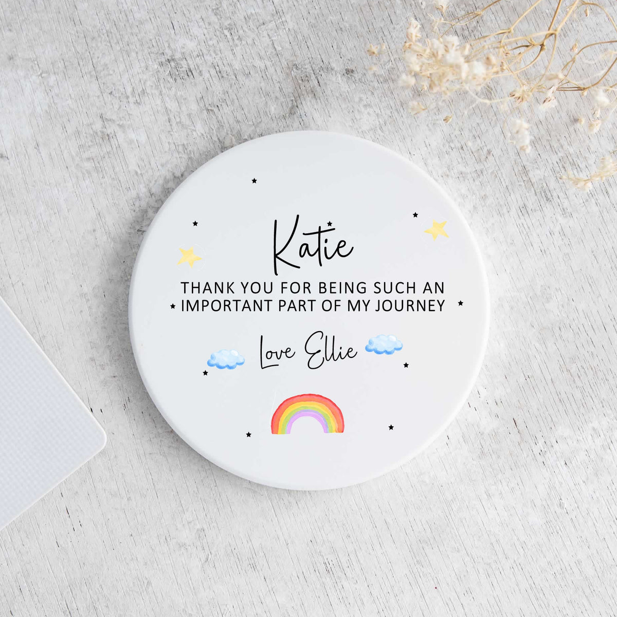 A round white coaster with personalized black text and small colorful illustrations, photographed from above on a light gray textured surface with part of a laptop and dried flowers visible.