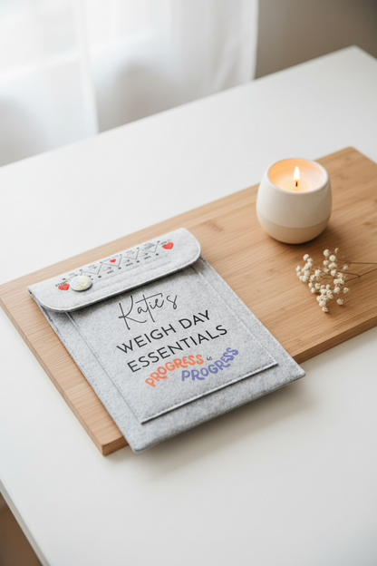A gray felt weight loss tracker pouch with a personalized design reading 'Katie's WEIGH DAY ESSENTIALS' and 'PROGRESS is PROGRESS' in orange and blue, shown on a white desk with a keyboard and binder clip nearby.