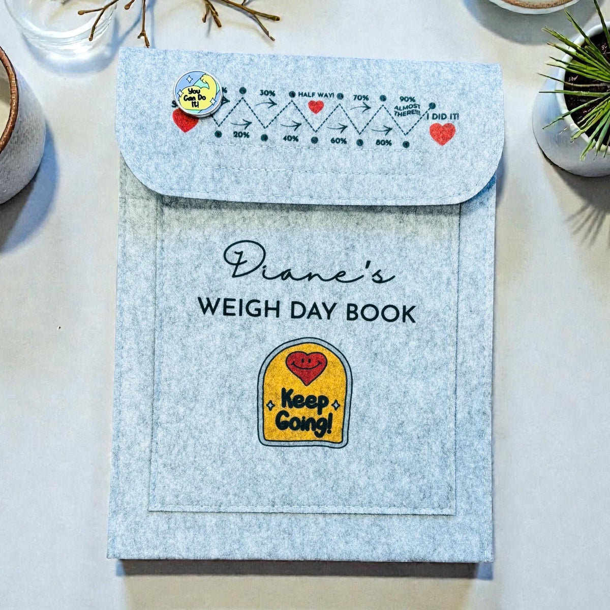 A light grey felt Weigh Day Book titled 'Diane's Weigh Day Book' with a flap cover, placed on a white surface with a ceramic mug, small plant, and decorative branches around it.