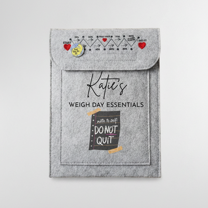 A gray felt pouch labeled 'Katie's WEIGH DAY ESSENTIALS' with a flap closure, printed weight-chart graphics, and a pocket reading 'write to self DO NOT QUIT', shown on a white desk with a keyboard corner and binder clip nearby.