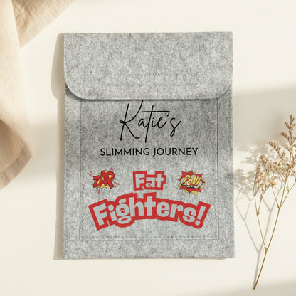 A gray felt sleeve with a flap closure, printed with the text 'Katie's Slimming Journey' and colorful retro-style graphics reading 'Fat Fighters!' in red, yellow, and white. The sleeve is photographed on a white desk with a keyboard corner and binder clips visible.