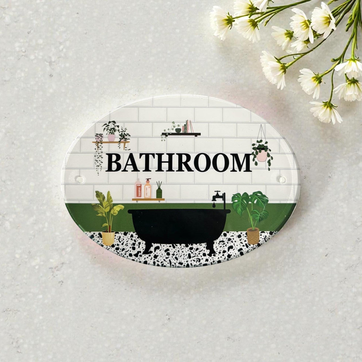 An oval bathroom-themed sign or plaque with a white subway tile background, black bathtub illustration, green plants, and the word “BATHROOM” in bold black letters. The sign is photographed on a light textured surface with small white flowers in the top-right corner.
