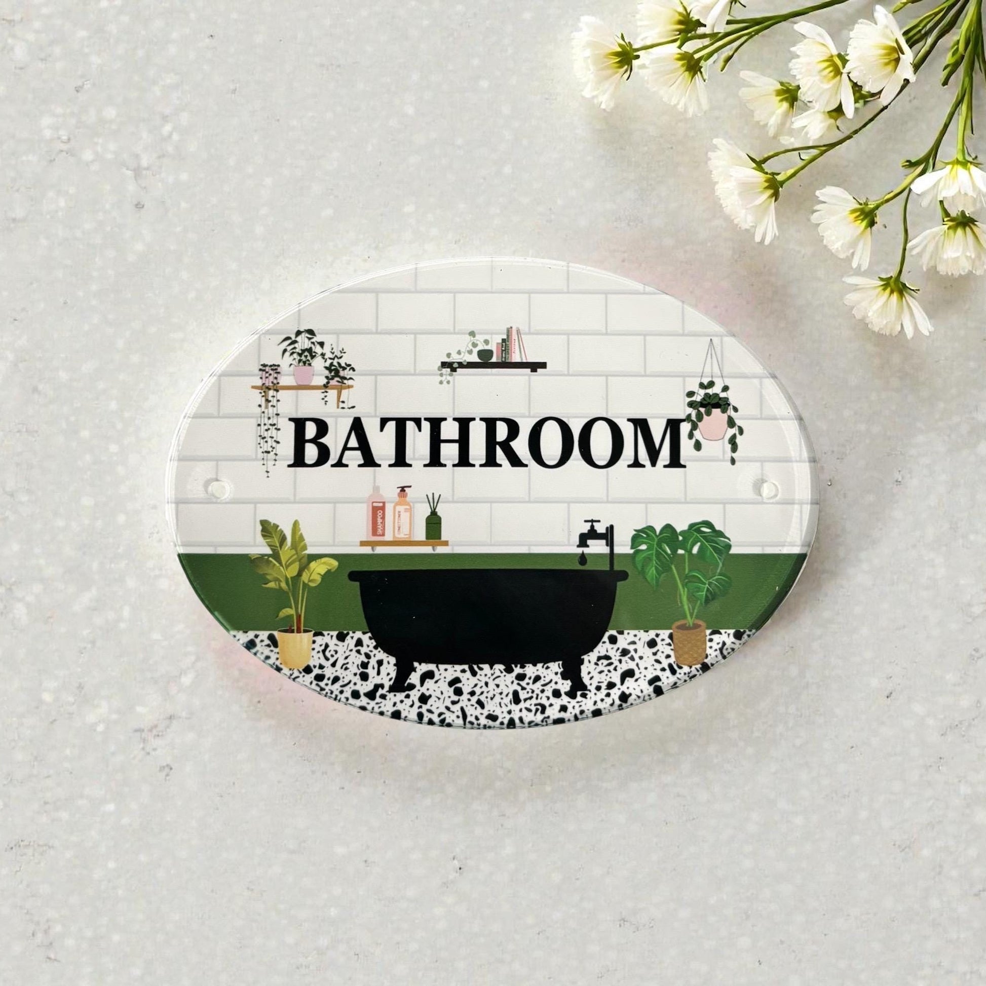 An oval bathroom-themed sign or plaque with a white subway tile background, black bathtub illustration, green plants, and the word “BATHROOM” in bold black letters. The sign is photographed on a light textured surface with small white flowers in the top-right corner.
