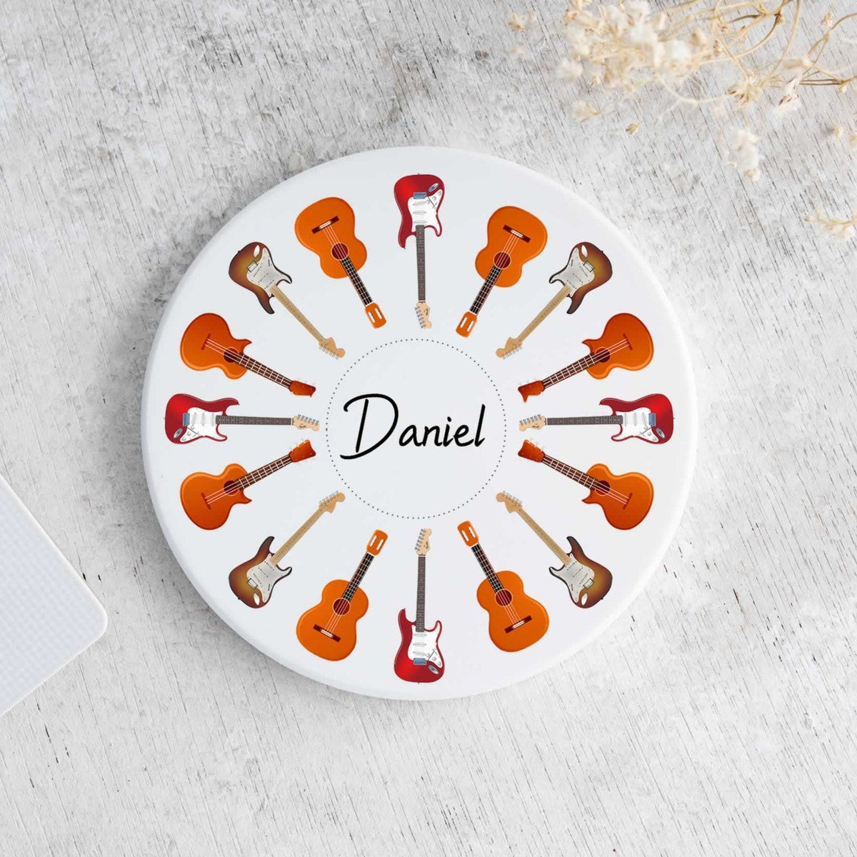 A round white coaster featuring a circular pattern of illustrated guitars in orange, brown, and red, with the name 'Daniel' printed in the center. The coaster is displayed on a light gray textured surface next to a laptop corner and dried flowers.