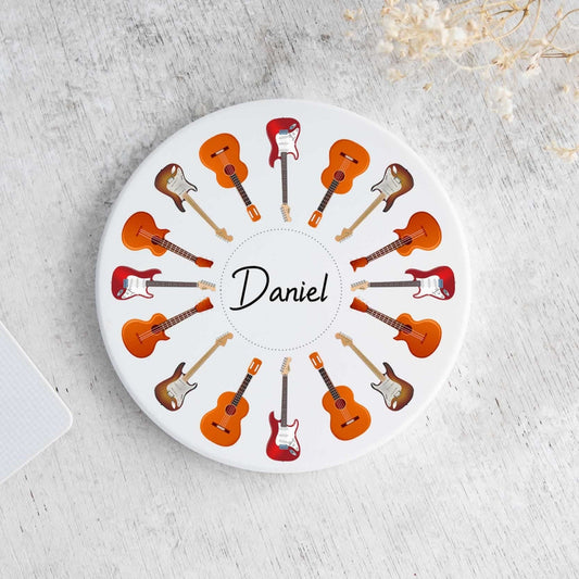 A round white coaster featuring a circular pattern of illustrated guitars in orange, brown, and red, with the name 'Daniel' printed in the center. The coaster is displayed on a light gray textured surface next to a laptop corner and dried flowers.